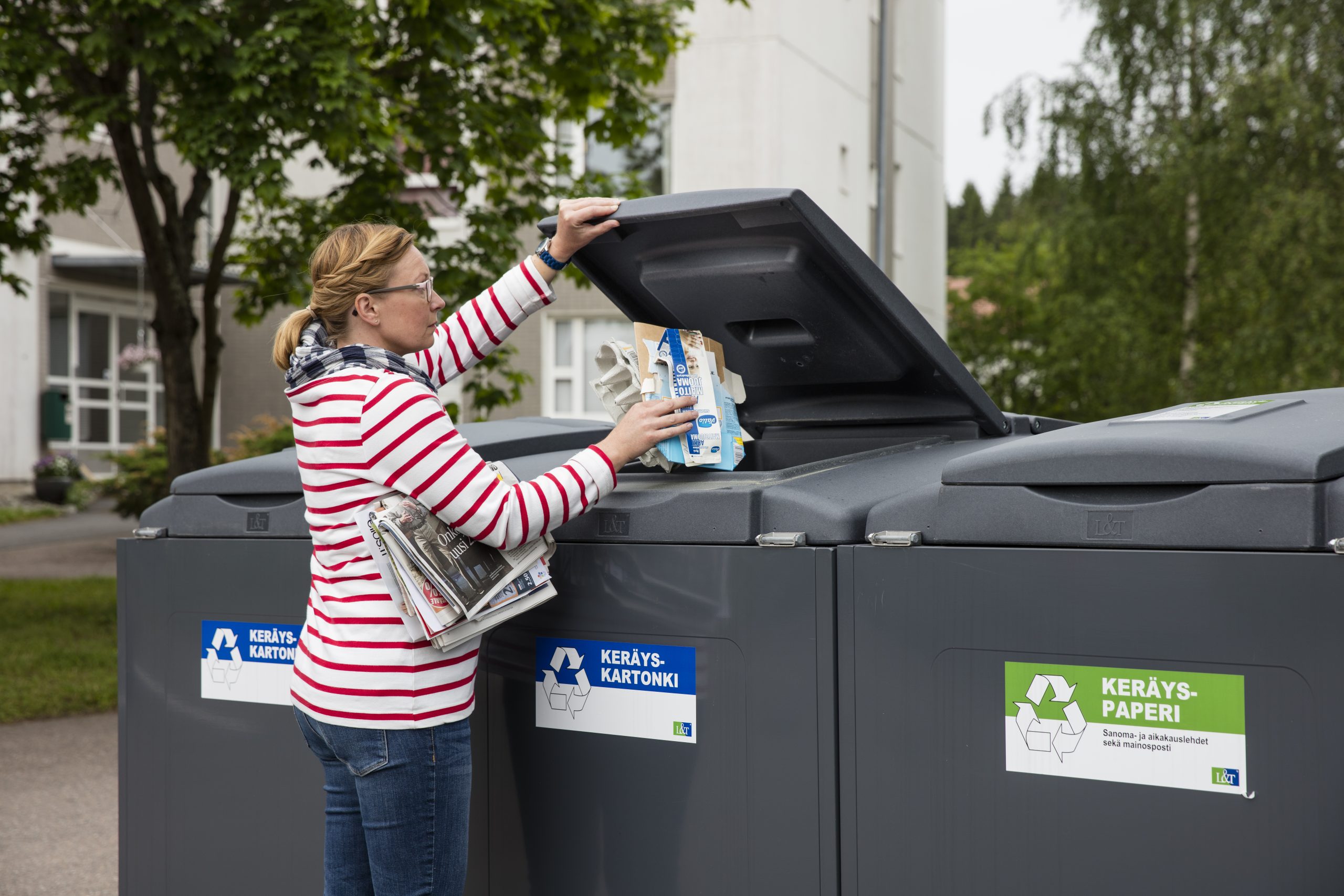 Nainen laittaa pahvia kartonkikeräykseen. Kainalossa on lehtiä seuraavaksi lehtilaariin laitettavaksi.