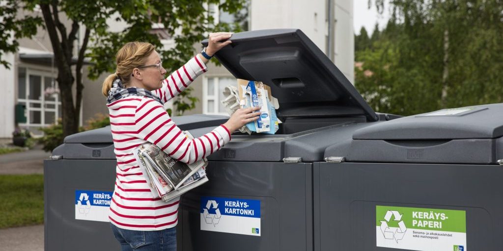 Nainen laittaa pahvia kartonkikeräykseen. Kainalossa on lehtiä seuraavaksi lehtilaariin laitettavaksi.